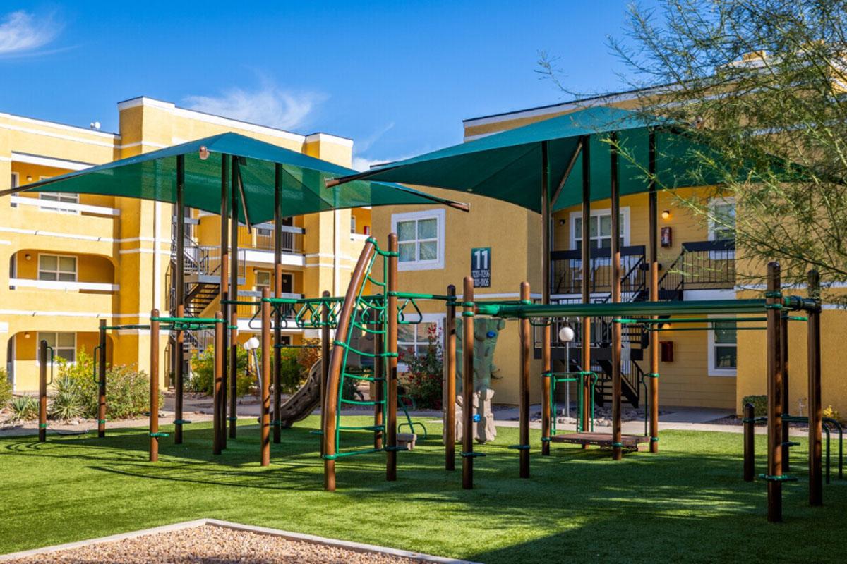 A colorful playground with various climbing structures and slides, surrounded by green grass. Large shade canopies provide coverage, with yellow apartment buildings in the background against a clear blue sky.