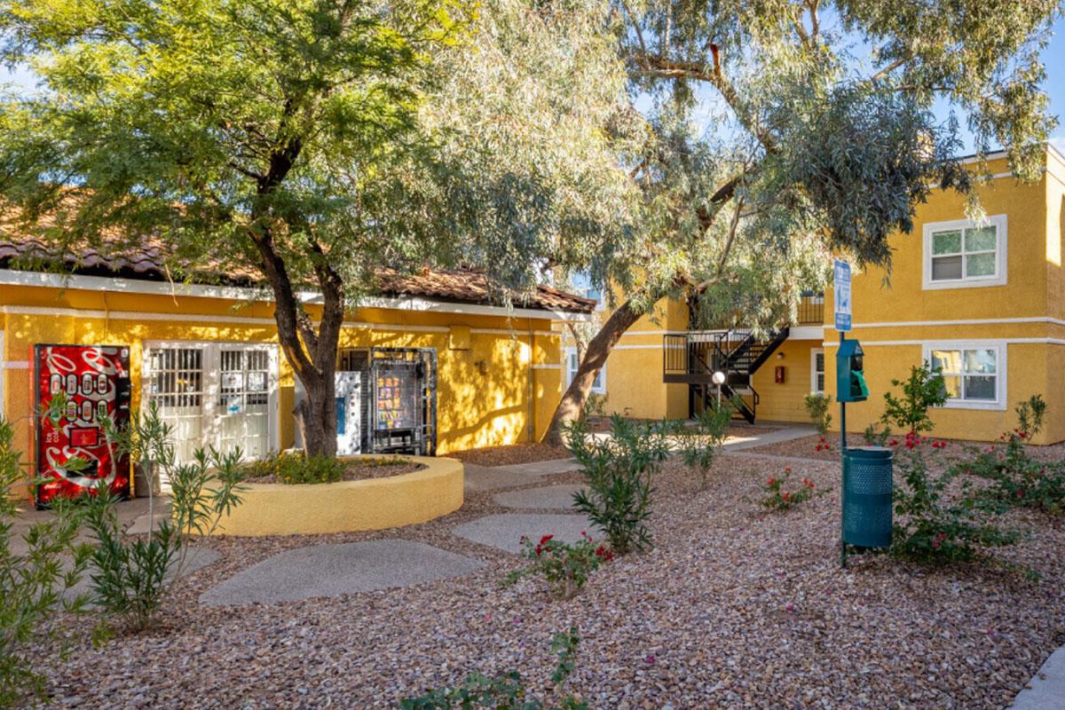 A view of a sunny courtyard featuring yellow buildings, surrounded by lush green trees and shrubs. There are decorative elements on the walls, a pathway made of gravel, and a small seating area. The architecture has a warm, inviting appearance typical of a residential complex.