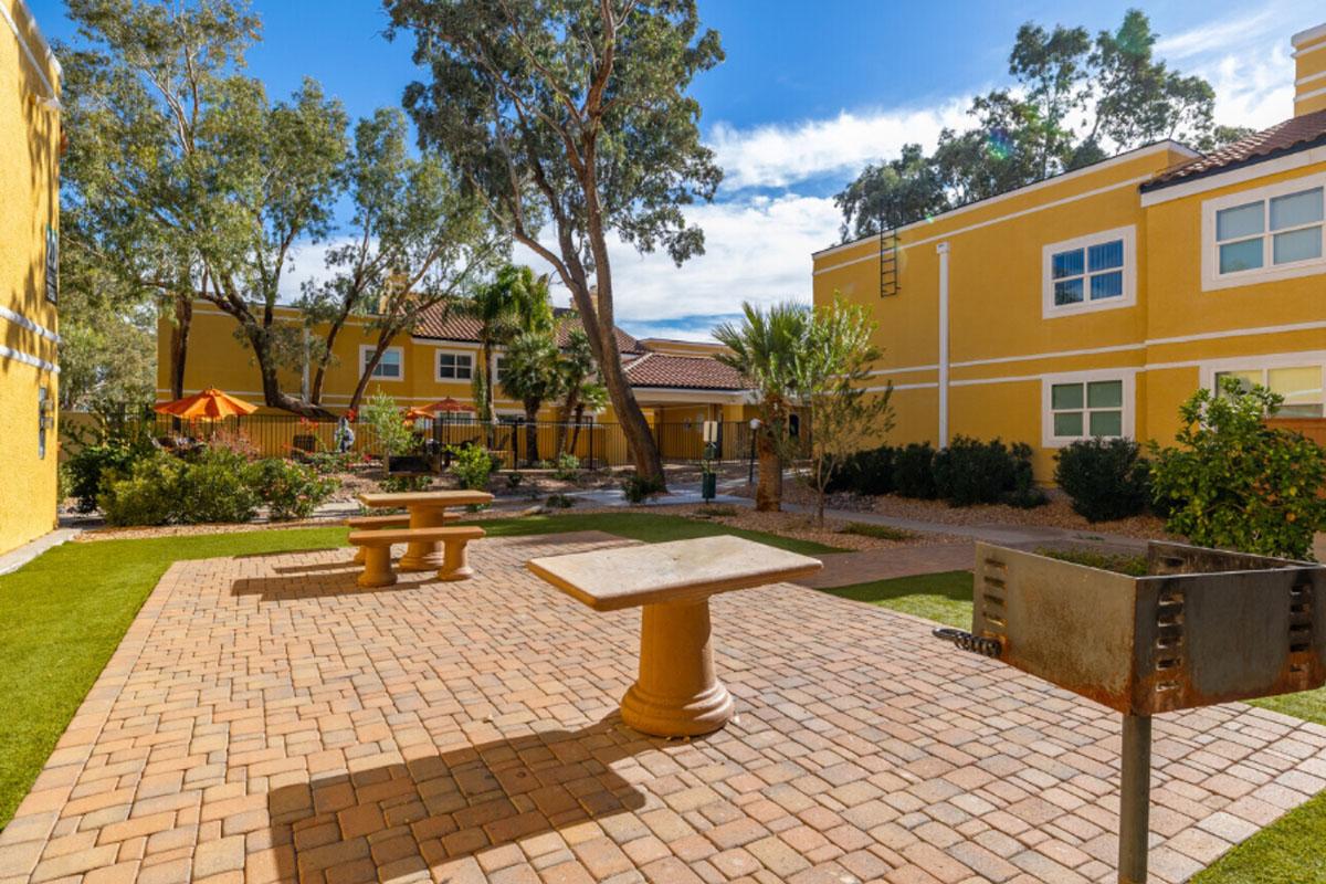 A landscaped outdoor area featuring a paved patio with a stone table in the foreground. Surrounding the patio are trees and shrubs, with bright yellow buildings in the background. There are umbrellas and seating areas visible, creating a welcoming atmosphere for relaxation and gatherings.