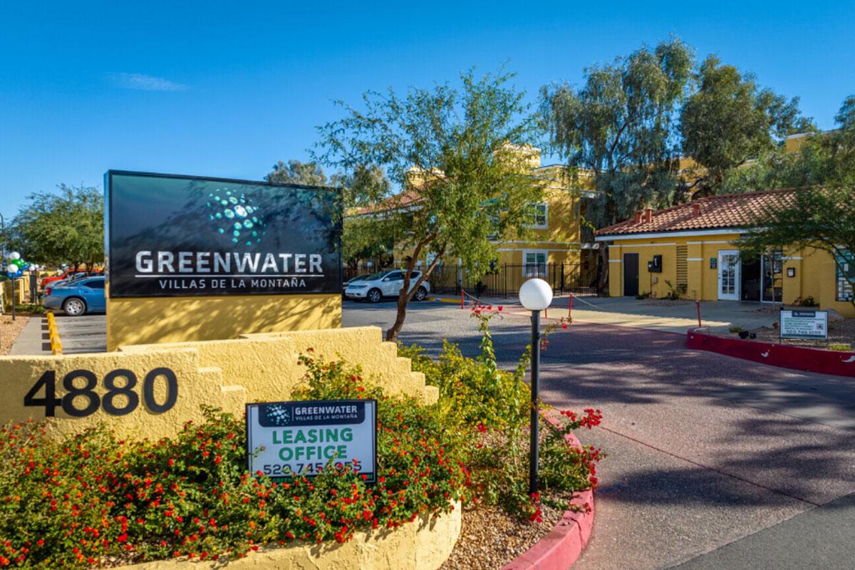 Signage for Greenwater Villas de la Montaña, featuring the address 4880. The entrance includes well-maintained landscaping with flowers, a prominent leasing office sign, and a view of the surrounding buildings and parking area under clear blue skies.