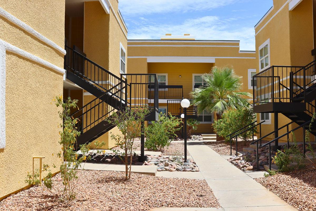 View of a bright courtyard surrounded by yellow apartment buildings. There are two staircases leading to second-floor entrances, lush greenery including a palm tree, and decorative rock landscaping. The sky is clear blue, adding to the vibrant atmosphere.