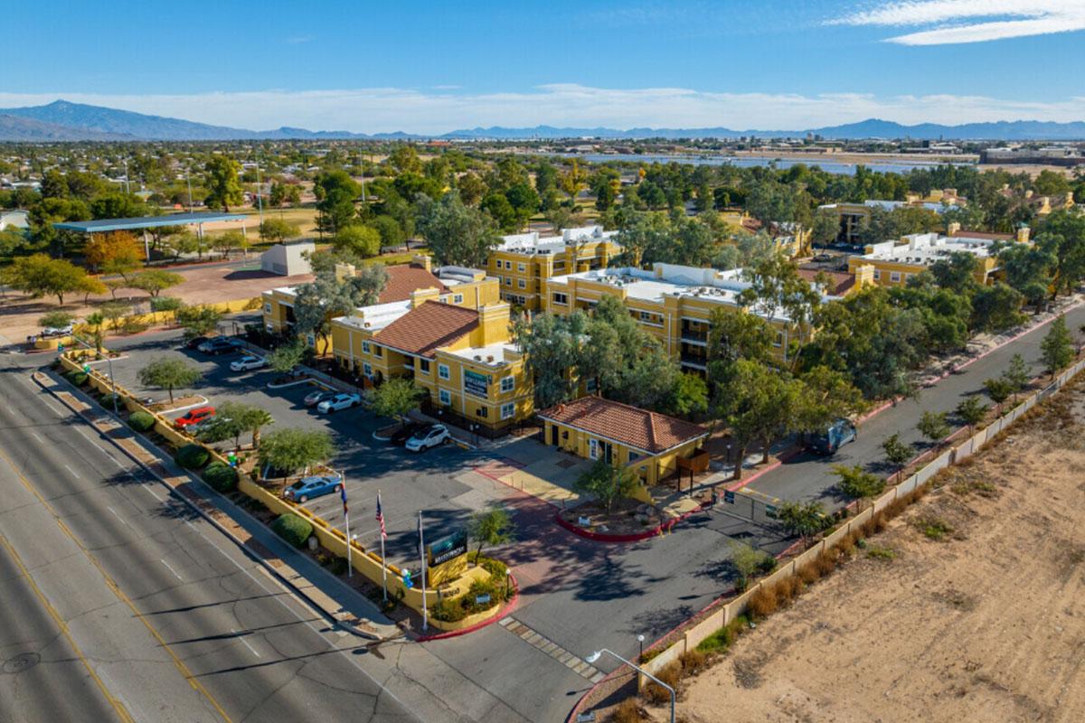 Aerial view of a yellow building complex surrounded by trees and parking spaces, with a road in the foreground. The landscape includes distant mountains and a clear blue sky, showcasing a suburban setting.