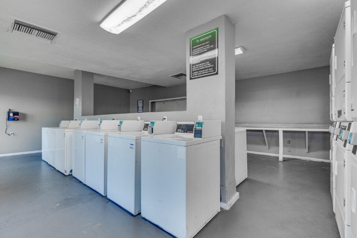 A clean, well-lit laundry room featuring several white washing machines and dryers lined up against a gray wall. The space has a simple, modern design with overhead lighting and a notice board visible on the wall.