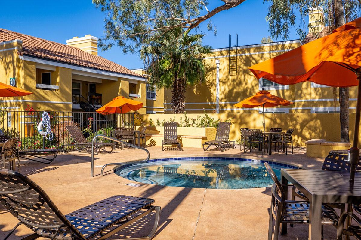 A sunny outdoor pool area featuring a hot tub surrounded by lounge chairs and orange umbrellas. There are landscaped plants in the background, with a yellow building and clear blue sky enhancing the relaxing atmosphere.