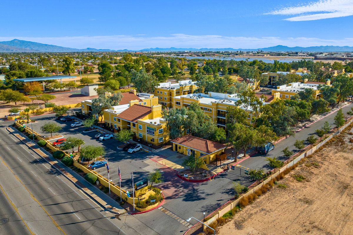 Aerial view of a yellow multi-story building complex surrounded by trees and parking lots, with a road in the foreground. In the background, mountains are visible under a clear blue sky. The scene captures a sunny day in a suburban area.