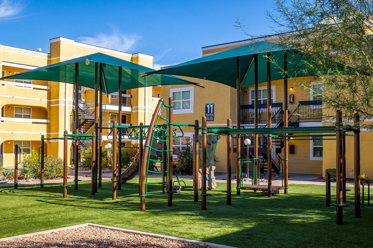 A children's playground with climbing structures and shaded areas, surrounded by green grass and apartment buildings. The playground features various play equipment designed for children, set against a bright blue sky.