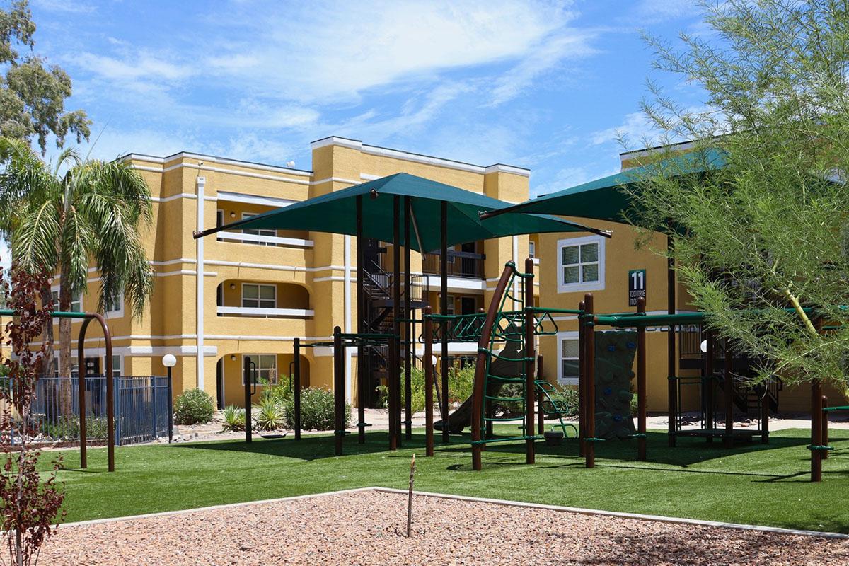 A sunny playground area with green grass, featuring a climbing structure and shaded play areas under large canopies. In the background, there are yellow apartment buildings with balconies and palm trees, set against a clear blue sky.
