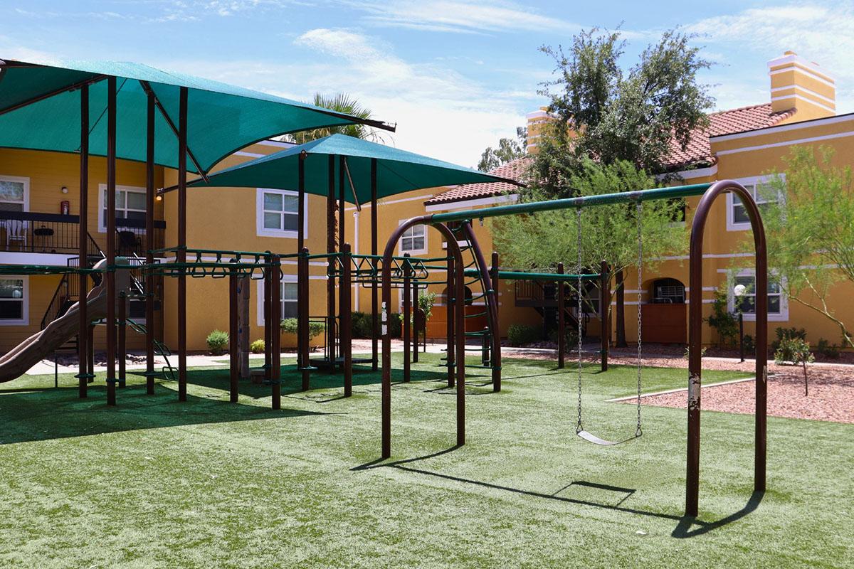 A playground featuring green grass, shaded areas with large canopies, and a set of swings. In the background, there are yellow apartment buildings surrounded by trees and shrubs, creating a bright and inviting outdoor space for children to play.