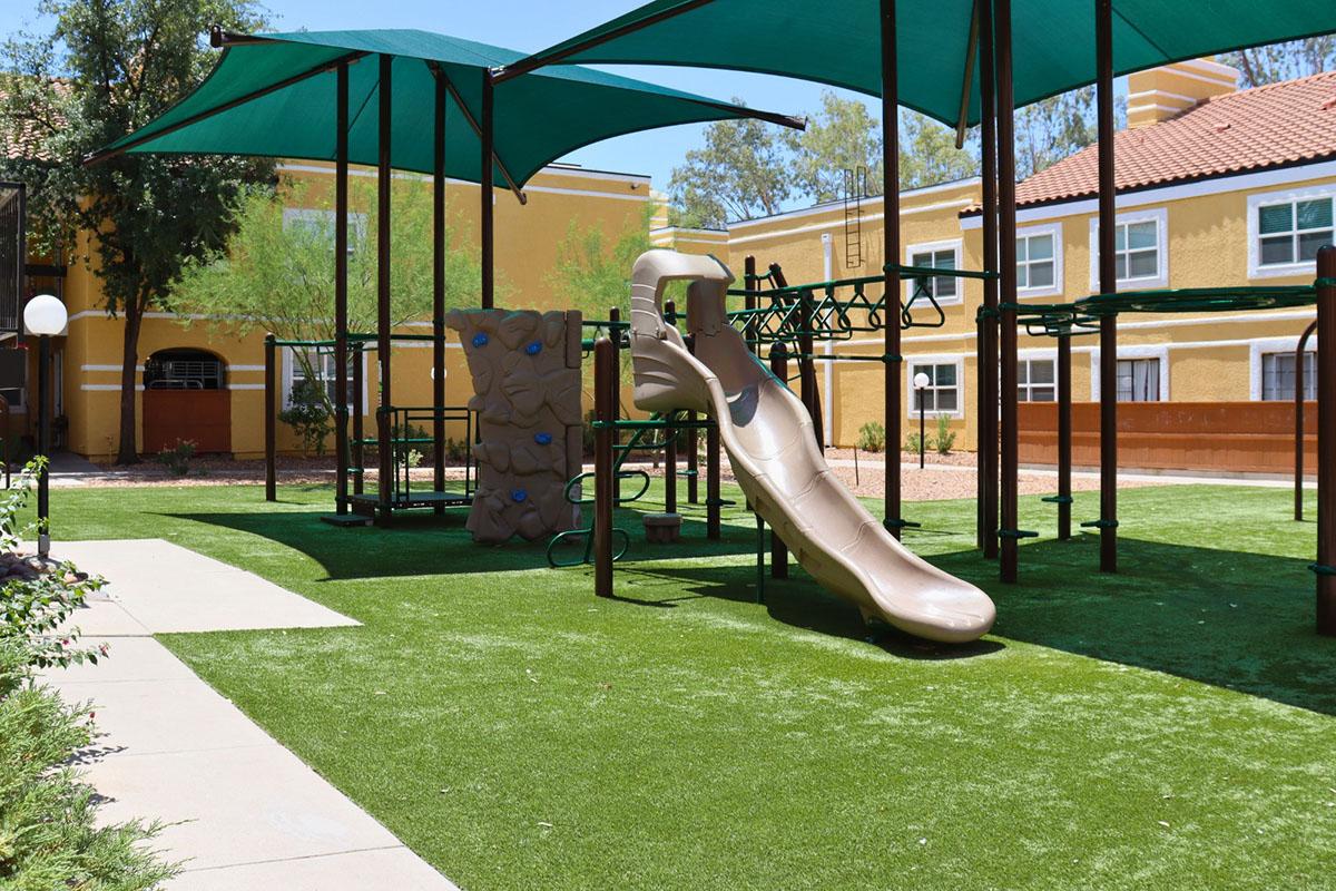 A playground scene featuring a large brown slide and a climbing structure on green artificial grass. The area is shaded by large tarps, with nearby residential buildings in the background. The setting is bright and inviting, suitable for children to play.