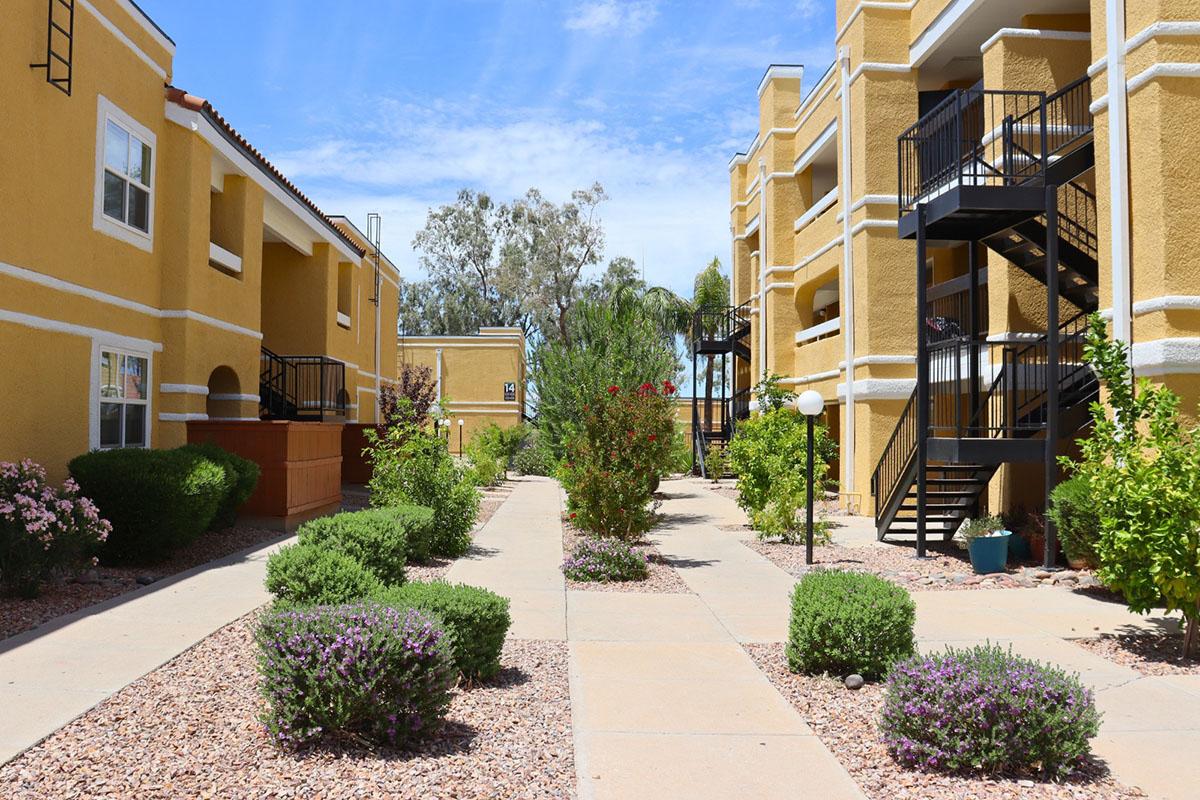 A sunny view of an apartment complex featuring two buildings with yellow exteriors. The pathway is lined with shrubs and flowers, with green bushes on either side. Black metal staircases are visible, and there are trees in the background, creating a pleasant outdoor environment.