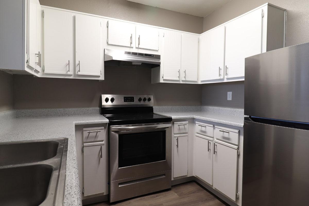 Modern kitchen featuring white cabinetry, stainless steel appliances including an oven and refrigerator, and a gray countertop. The space is well-lit with a neutral color scheme and has a double sink under the counter. The layout is efficient, ideal for cooking and food preparation.