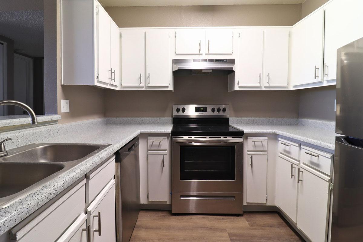 A modern kitchen with white cabinets, a stainless steel refrigerator, a dishwasher, and a black stove and oven. The countertops are light speckled, and the space is well-lit, creating a clean and inviting atmosphere. The flooring is wood-like, enhancing the contemporary feel of the kitchen.