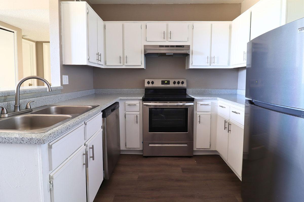 A modern kitchen featuring white cabinetry, stainless steel appliances, including a stove, refrigerator, and dishwasher. The counter has a speckled design, and there are dual sinks under a window. The floor is a wood laminate, enhancing the contemporary feel of the space.