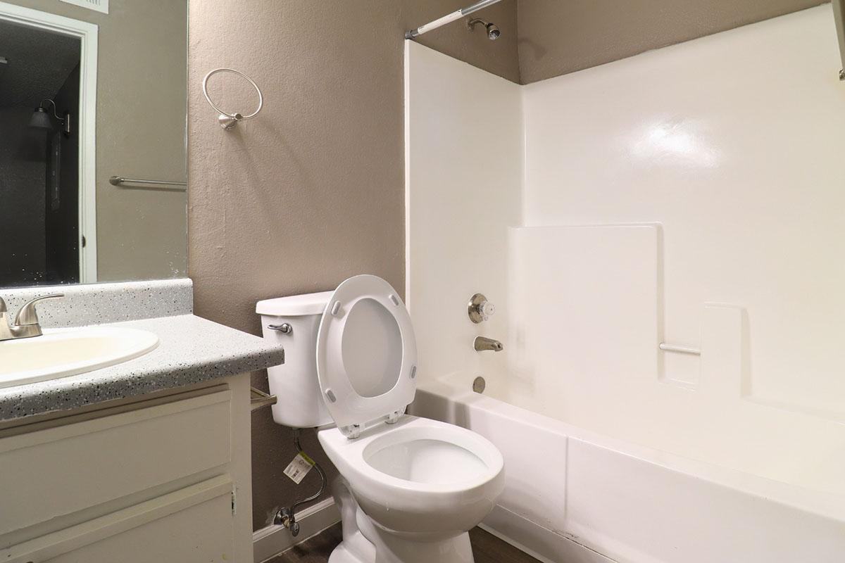 A small bathroom featuring a white bathtub with a shower, a toilet, and a sink with a granite countertop. The walls are painted a neutral color, and the floor appears to be a wood or laminate surface. A towel bar is mounted on the wall beside the sink.