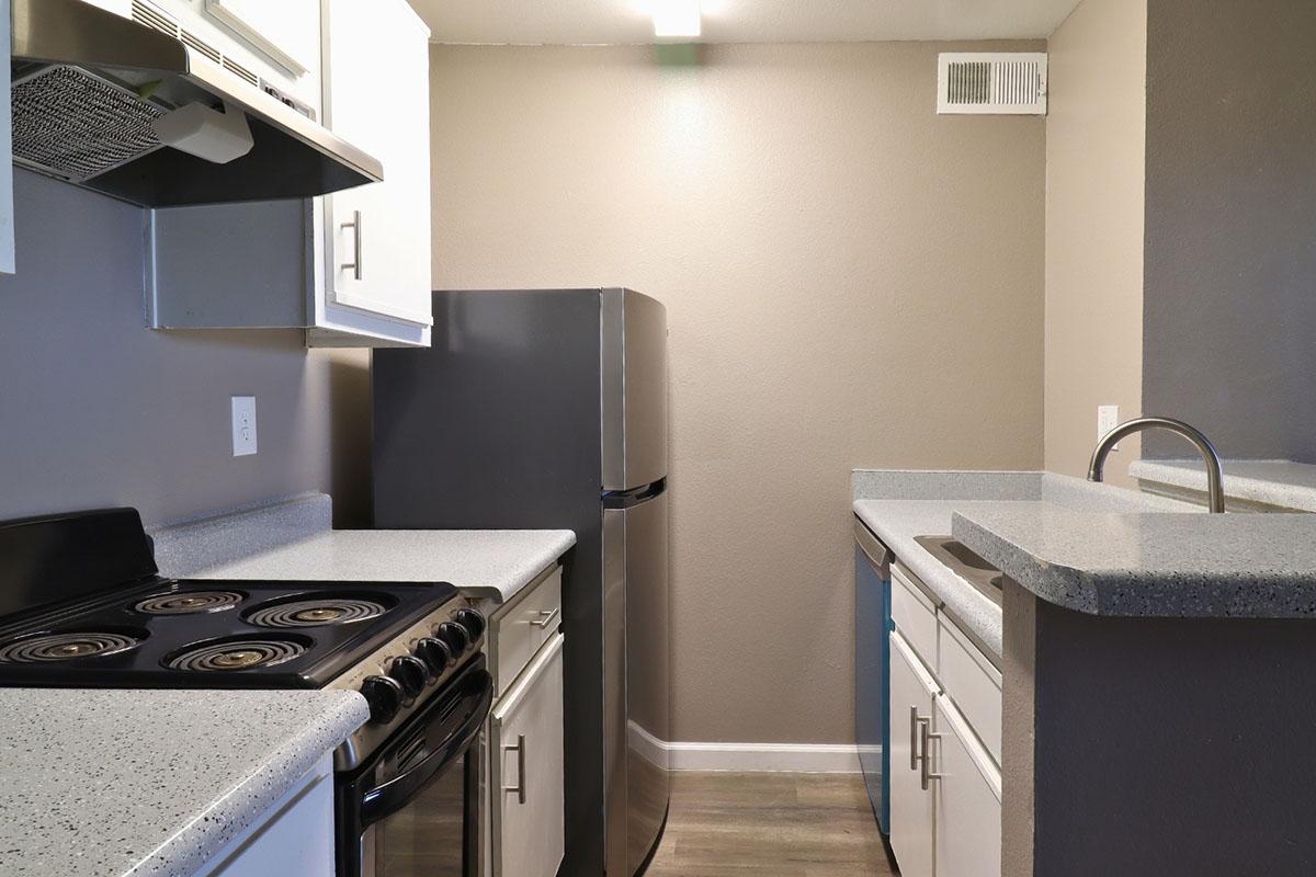 A modern kitchen featuring gray walls, a refrigerator, a stove, and white cabinetry. The countertops are light gray with a speckled pattern. The kitchen has a narrow layout with a sink positioned in front of a window. Adequate lighting is provided, enhancing the clean and minimalist appearance.