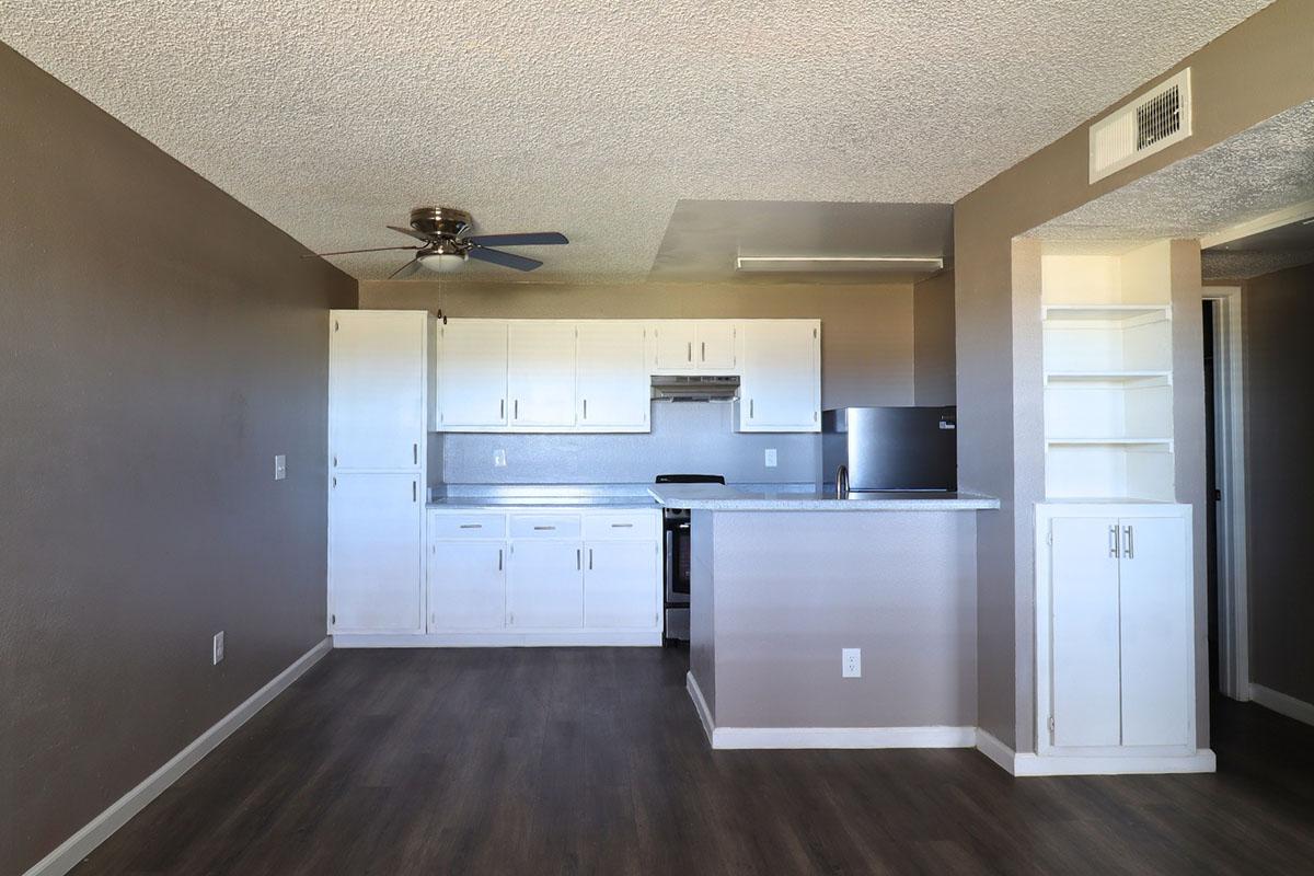 A modern kitchen and living area with gray walls and wood-like flooring. The kitchen features white cabinets, a stove, and a small countertop. A ceiling fan is visible, along with built-in shelves. The space is well-lit and open, creating a cozy atmosphere.