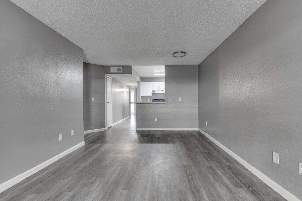 A spacious, modern living area with gray walls and laminate flooring. The room features an open layout leading to a kitchen with white cabinetry, visible in the background. Natural light illuminates the space, creating an inviting atmosphere.