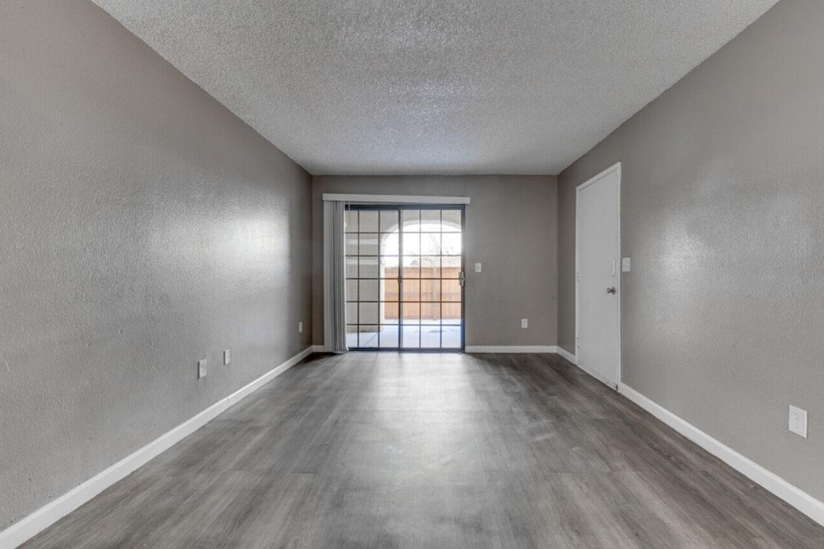 A spacious empty living room with light gray walls and a wood-like floor. Large window with a sliding door leads to a patio, allowing natural light to fill the space. The room features a simple, minimalistic design with no furniture, creating an open and airy atmosphere.