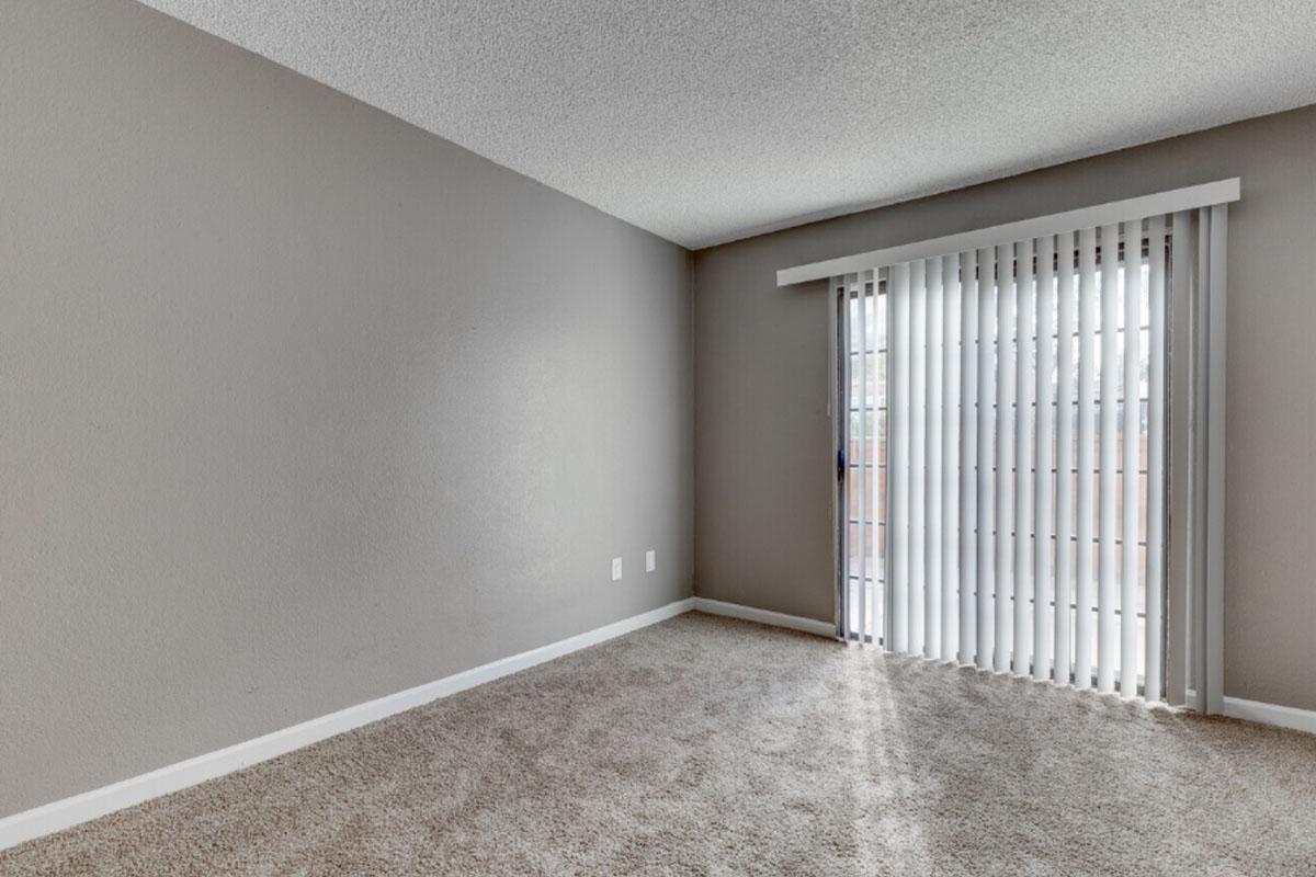 Empty room with light gray walls and a light carpet. A sliding glass door with vertical blinds allows natural light to enter. The space is uncluttered, featuring two electrical outlets on the wall and a window that opens to an outdoor area. No furniture or decorations are visible.