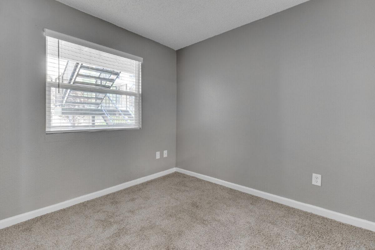 A small, empty room with light gray walls and a carpeted floor. There is a window with white blinds letting in natural light, revealing a view of an exterior structure. The room has minimal decor, with electrical outlets on the walls and no furniture present.