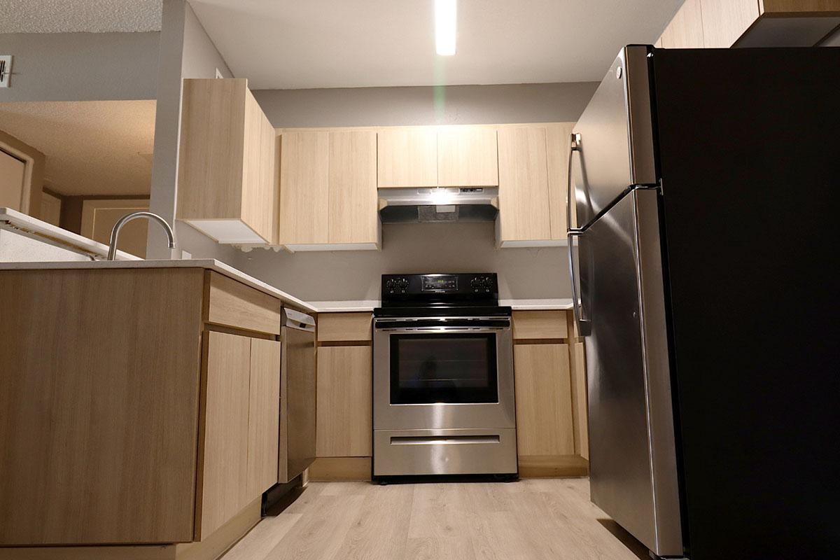 Modern kitchen featuring light wood cabinetry, stainless steel appliances, including a stove and refrigerator. The countertop is clear, and the flooring is light-colored vinyl or laminate. Bright overhead lighting highlights the spacious design. Overall, the kitchen has a clean and contemporary look.