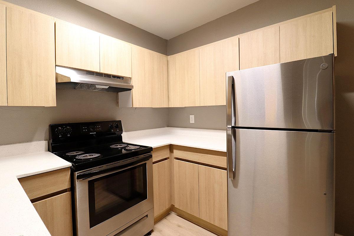 Modern kitchen featuring light wood cabinetry, a stainless steel refrigerator, and an electric stove with an overhead exhaust fan. The countertop is white, and the walls are painted in a neutral gray. The design emphasizes a clean and open feel, ideal for cooking and entertaining.