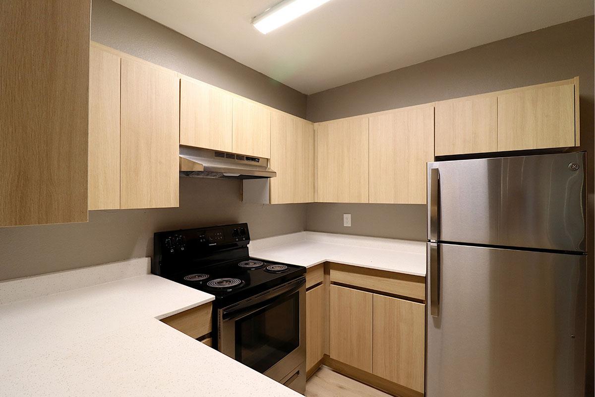 Modern kitchen featuring light wood cabinetry, stainless steel refrigerator, and a black electric stove with an oven. The countertops are white and the walls are painted a muted gray. The space is well-lit with overhead lighting.