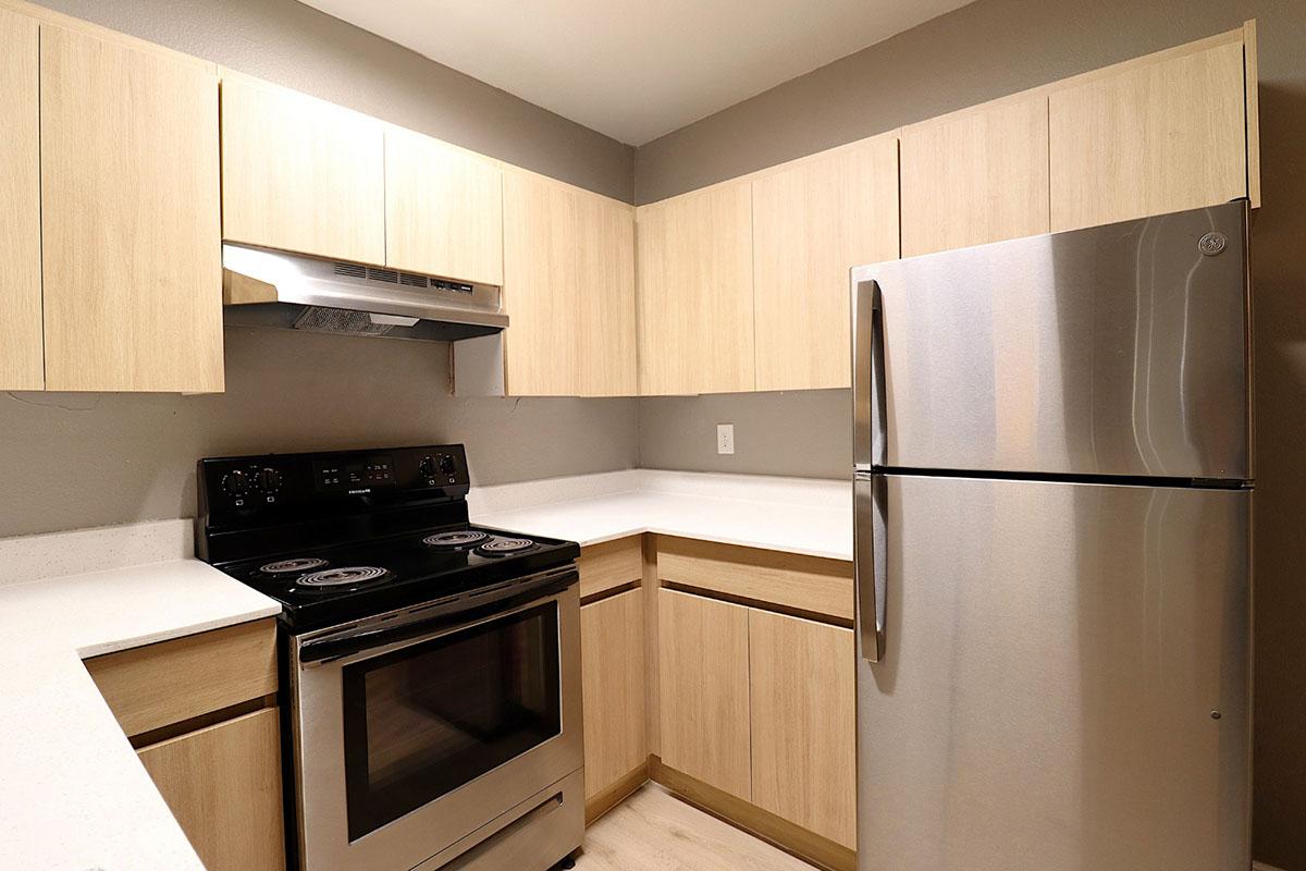 A modern kitchen featuring light wood cabinetry, a stainless steel refrigerator, and an electric stove with an oven. The countertop is white with a smooth finish, and the walls are painted a neutral gray. The overall design is sleek and contemporary, suitable for a small apartment or modern home.