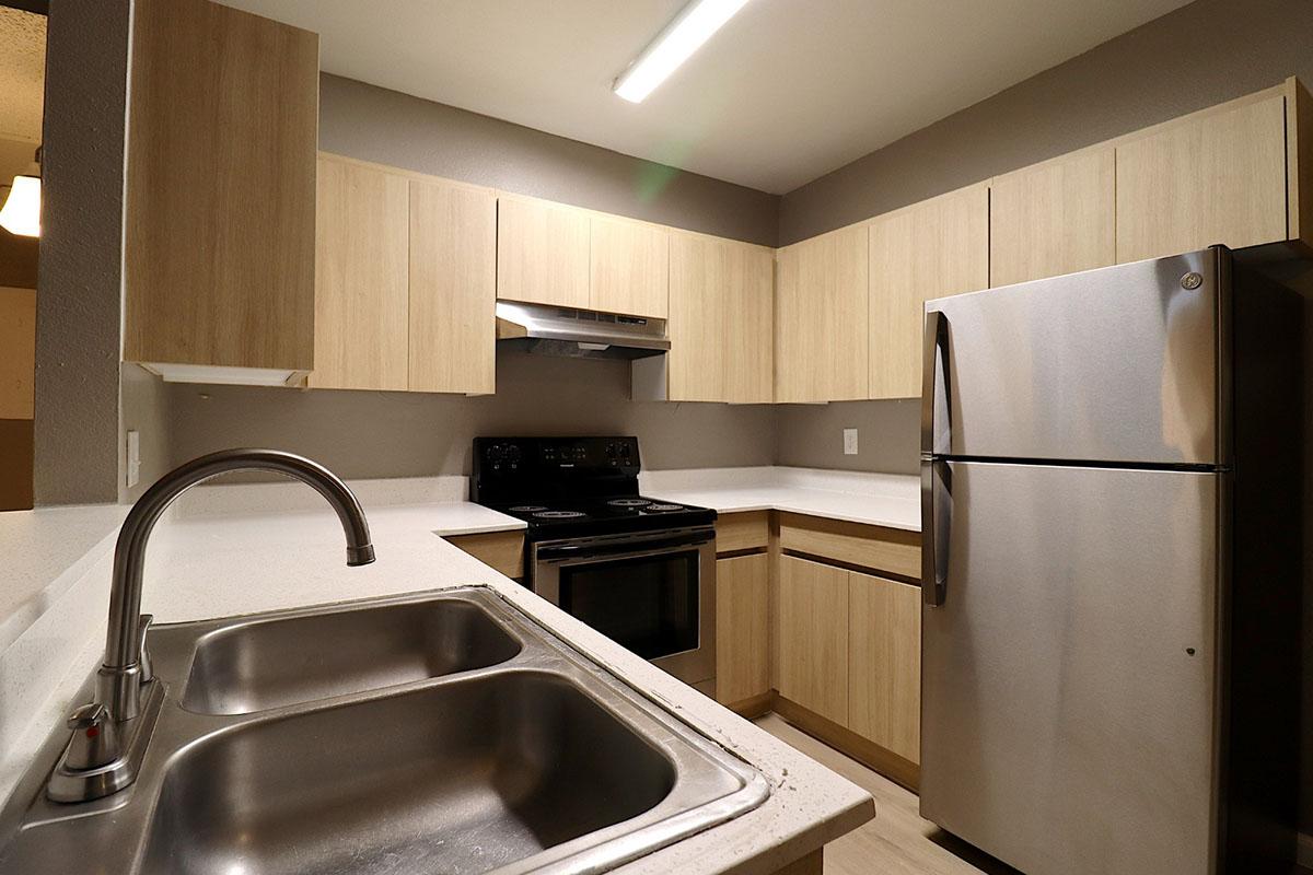 Modern kitchen featuring light wood cabinetry, stainless steel refrigerator, black stove, and a double sink. The space has a neutral color palette with a simple, clean design and overhead lighting.