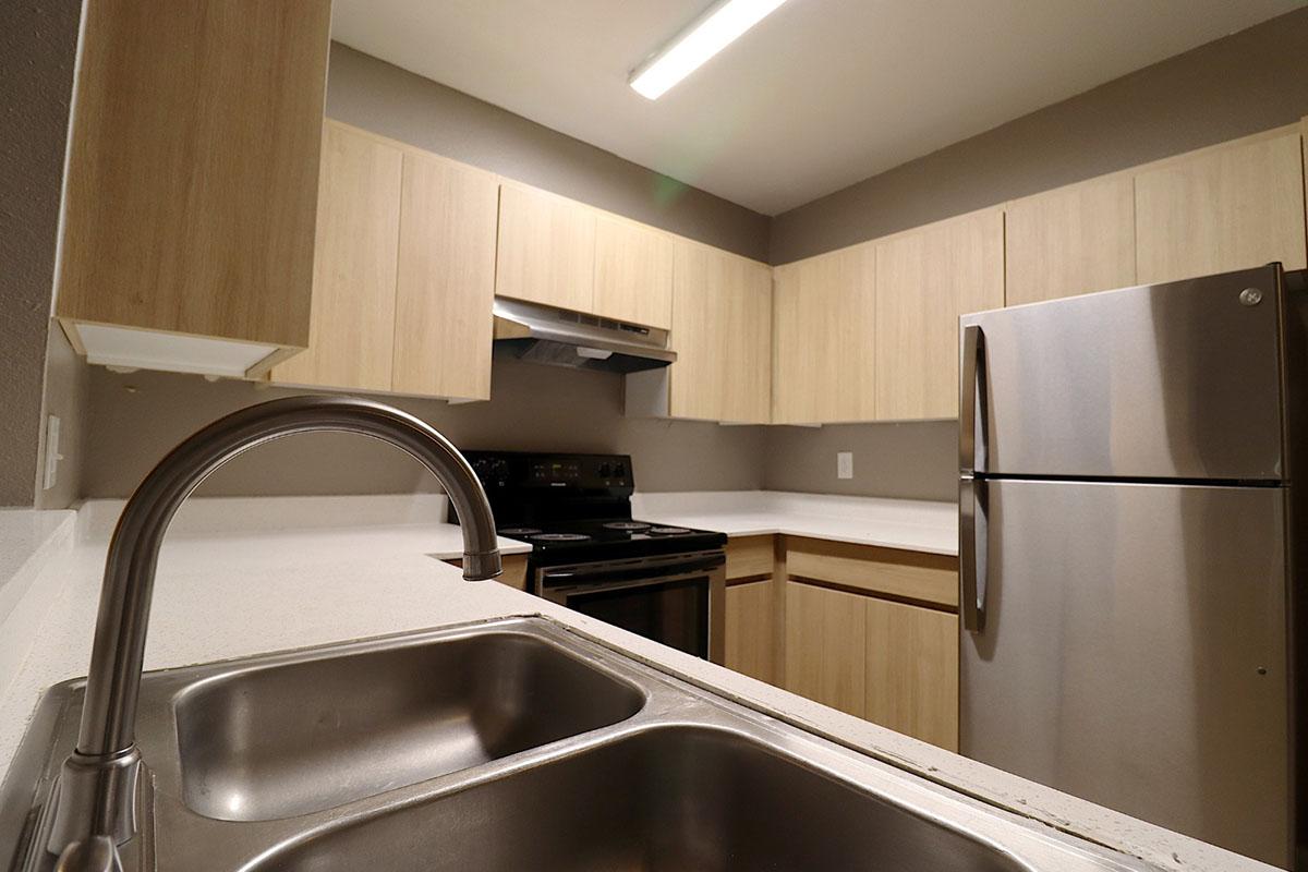 Modern kitchen with light wood cabinetry, stainless steel appliances including a fridge and stove, and a double sink. The countertops are white and the lighting features a fluorescent strip. The walls are painted a neutral color, creating a bright and clean atmosphere.