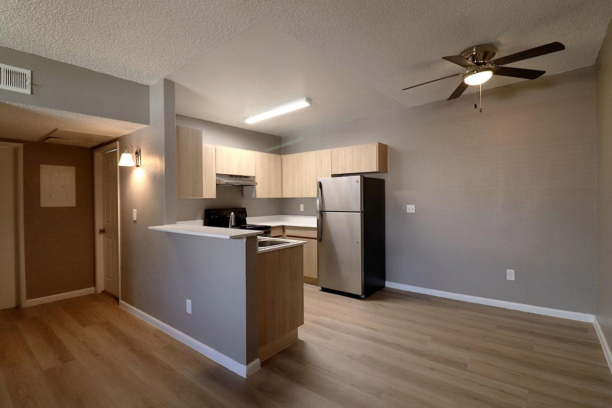 A modern kitchen and living area in an apartment, featuring light wood cabinetry, stainless steel appliances, and a ceiling fan. The space has a light gray color scheme with laminate flooring and a small kitchen island. Wall lights provide additional illumination.