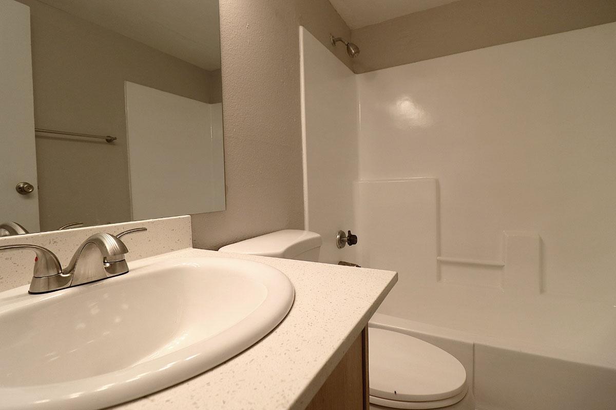 A bathroom featuring a white countertop with a sink and modern faucets, a white bathtub with a shower, and an adjacent toilet. The walls are painted in a neutral tone, and there is a mirror above the sink. The overall design is clean and minimalistic.