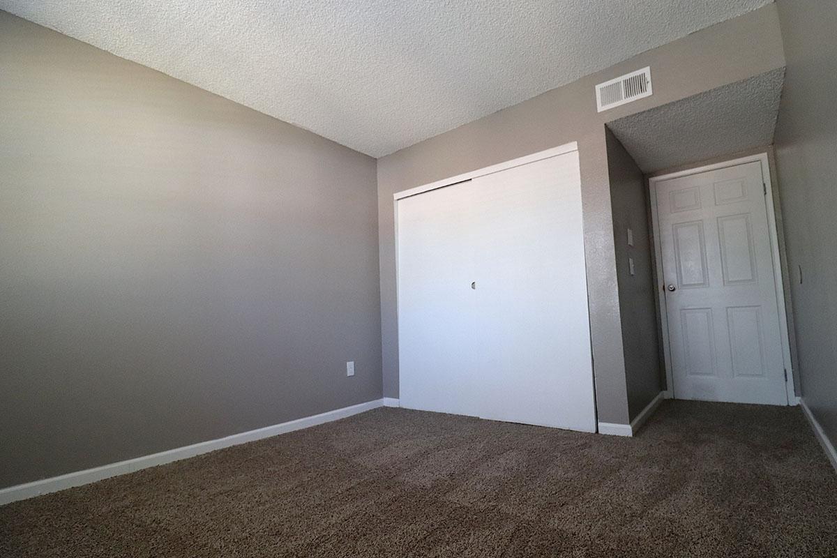 Empty room with gray walls and carpeting. There is a white closet door on the left and a closed door leading to another area on the right. Natural light is coming from the left, creating a bright but sparse atmosphere. The space is unfurnished, giving it a clean and minimalist look.