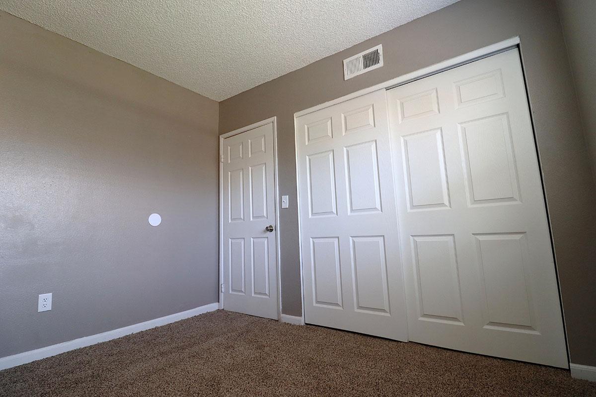 A view of a sparsely furnished room with light beige walls, a carpeted floor, and two white closet doors. There is a closed door on the left side of the image and a small circle on the wall, suggesting a light switch or outlet. The room has minimal decor, giving it a clean, open appearance.