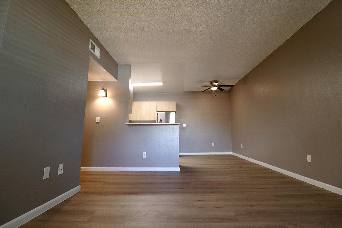 Spacious room with light gray walls, wood-style flooring, and a ceiling fan. A kitchen area is partially visible at the back, featuring light cabinets and appliances. The layout is open, creating a bright and airy atmosphere.