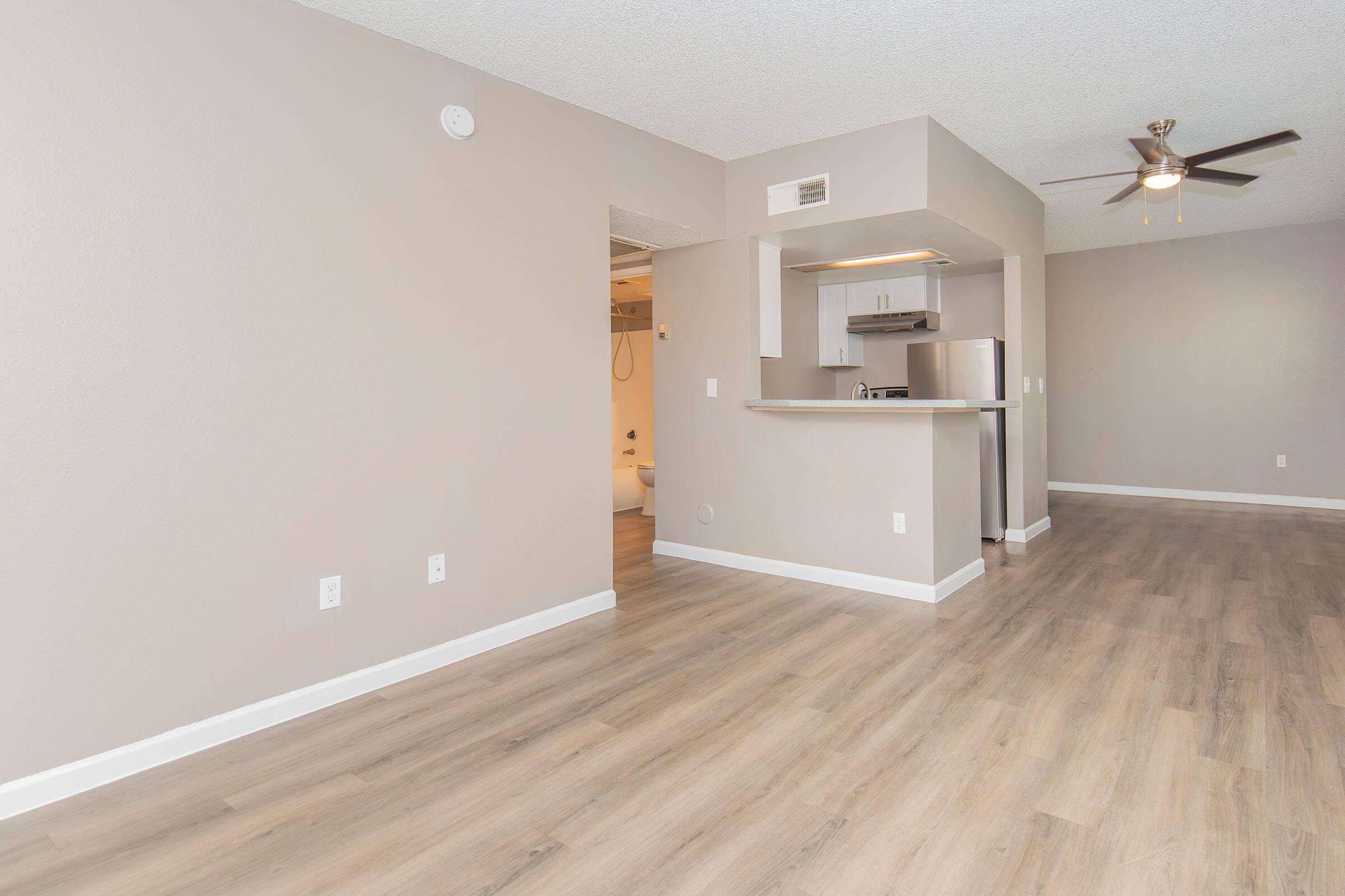 A spacious, modern apartment interior featuring light-colored walls, wooden flooring, and an open kitchen area. The kitchen includes stainless steel appliances and a bar counter. The living area has natural light, and the ceiling fan adds to the contemporary ambiance.