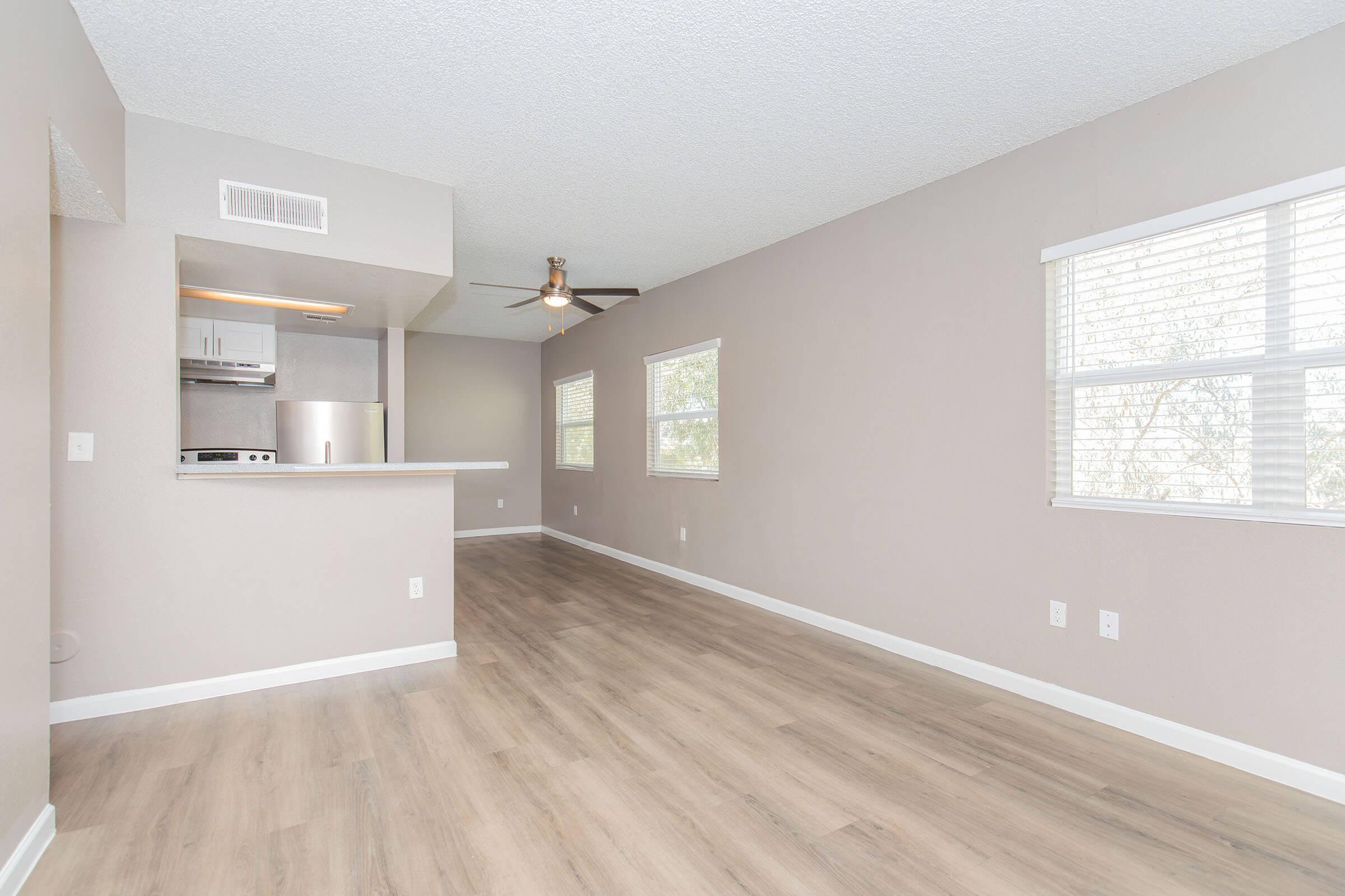A spacious indoor living area with light-colored walls and wood-style flooring. The room features a ceiling fan and multiple windows that allow natural light to flow in. An open kitchen layout is visible in the background, highlighting a bar counter and modern appliances.