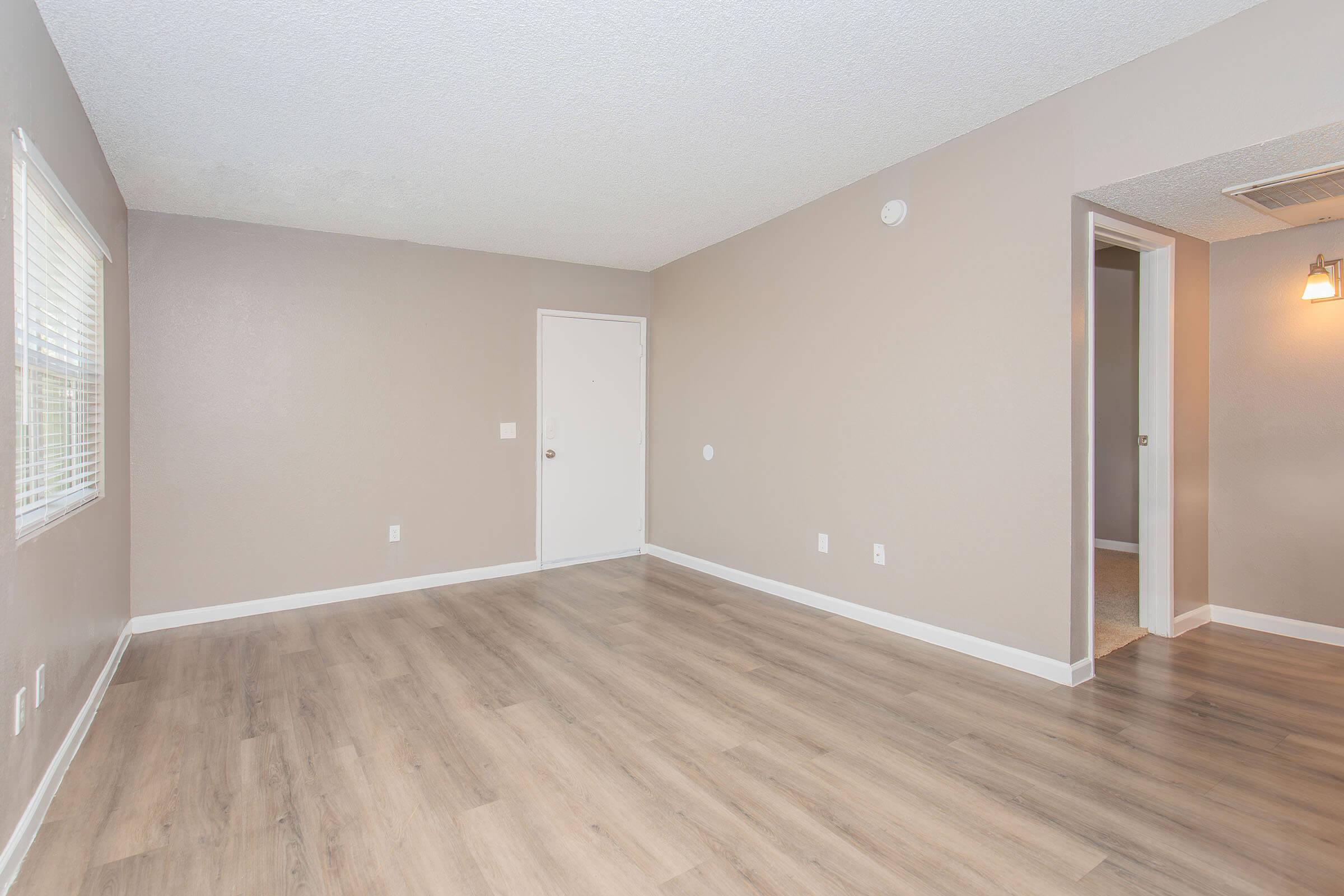 Empty room with light brown walls and a textured ceiling. The flooring is light wood. There is a closed door on the left and an open doorway leading to another room on the right. A window with blinds is visible on the front wall, allowing natural light to enter the space.