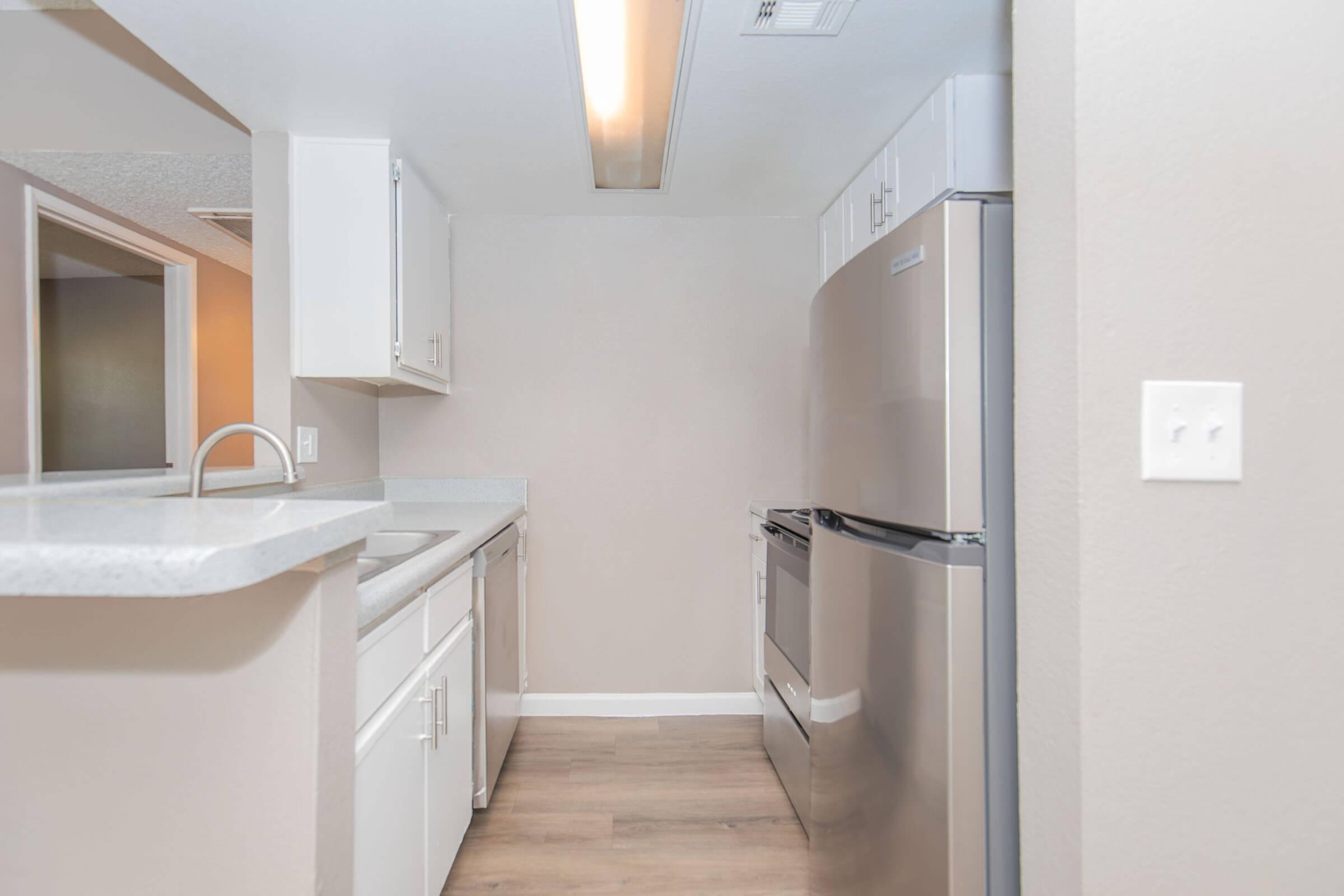 A modern kitchen featuring white cabinetry, a stainless steel refrigerator, and a smooth countertop. Natural light illuminates the space, highlighting the wooden flooring. The kitchen is designed with an open layout, providing a clear view into the adjacent room.