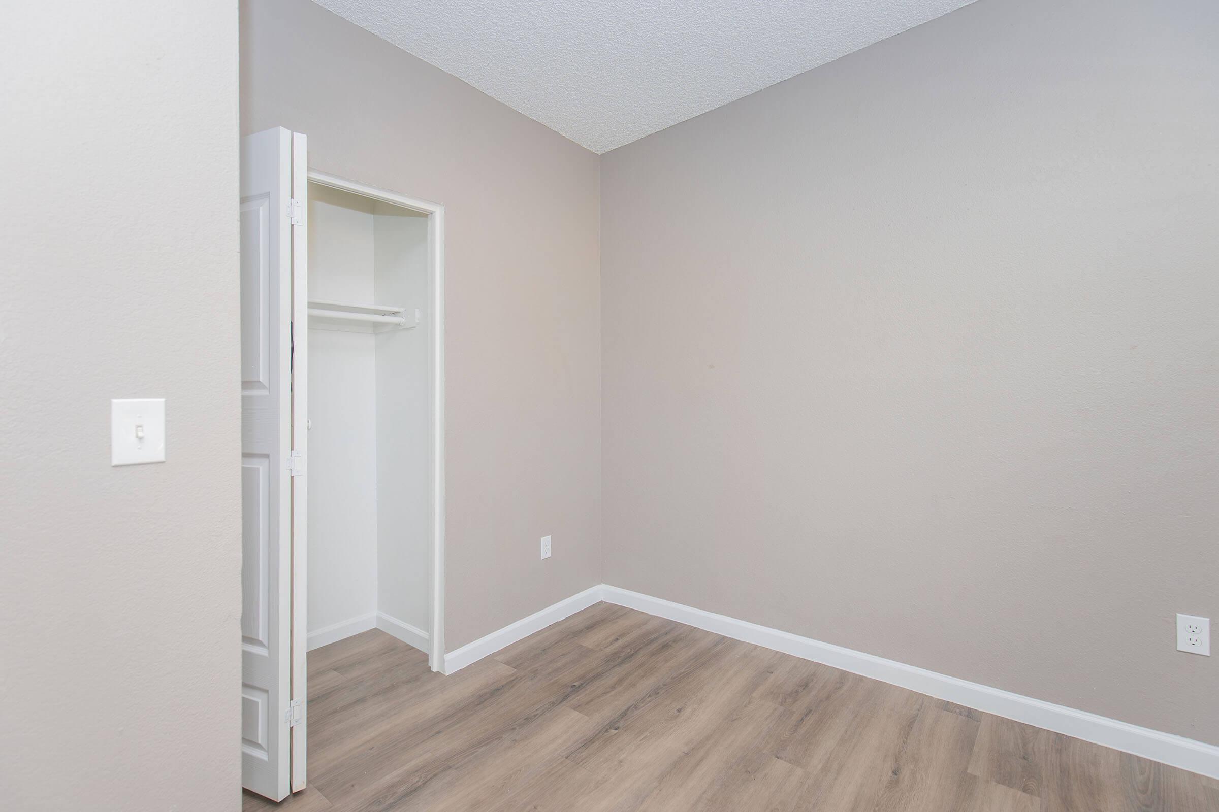 A sparsely furnished room featuring light gray walls, a wooden floor, and a closet with sliding doors. The room has no visible furniture, creating a minimalist appearance, with one corner showcasing the closet and the rest of the space empty.