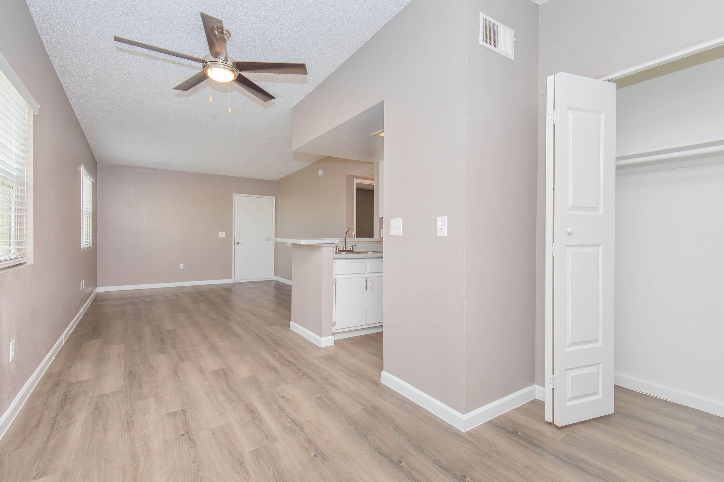 A spacious living area with light gray walls, a ceiling fan, and wood-style flooring. A doorway leads to a small kitchen area with a bar. An open closet with white doors is visible, and there are windows providing natural light. Overall, the space appears bright and inviting.