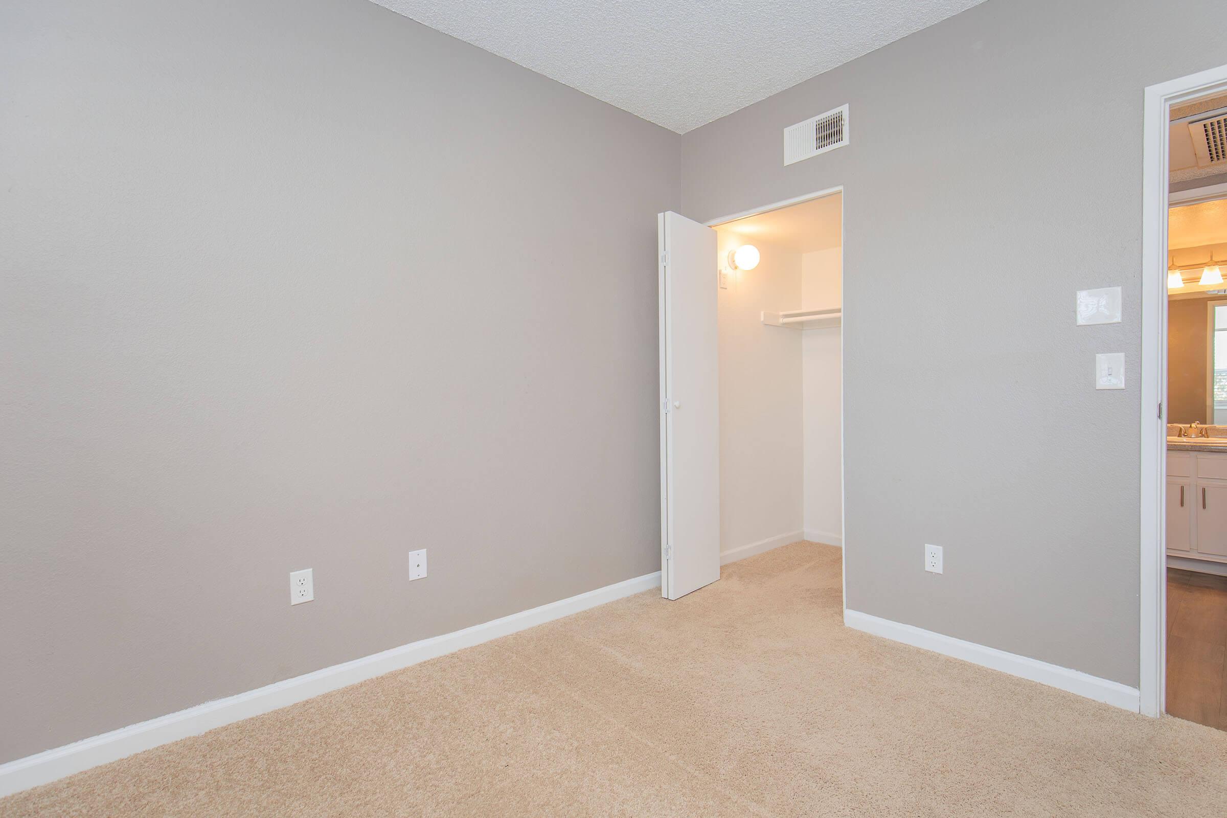 A partially empty room with gray walls and beige carpet. A white door leads to a closet on the left, while an open door on the right leads to another area featuring lighting. The room appears well-lit and spacious, with no furniture present.