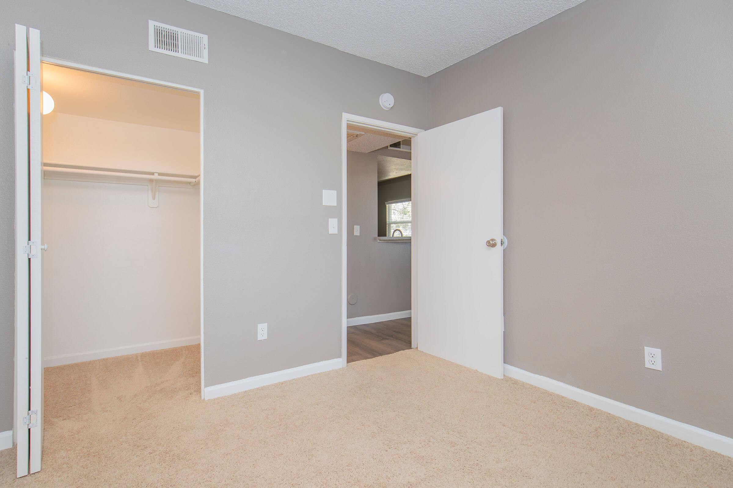 A well-lit, empty room featuring neutral gray walls and light-colored carpet. There are two doors: one leading to a closet and another possibly leading to another area. The window allows natural light, creating a spacious and airy ambiance. Ideal for a bedroom or office setup.
