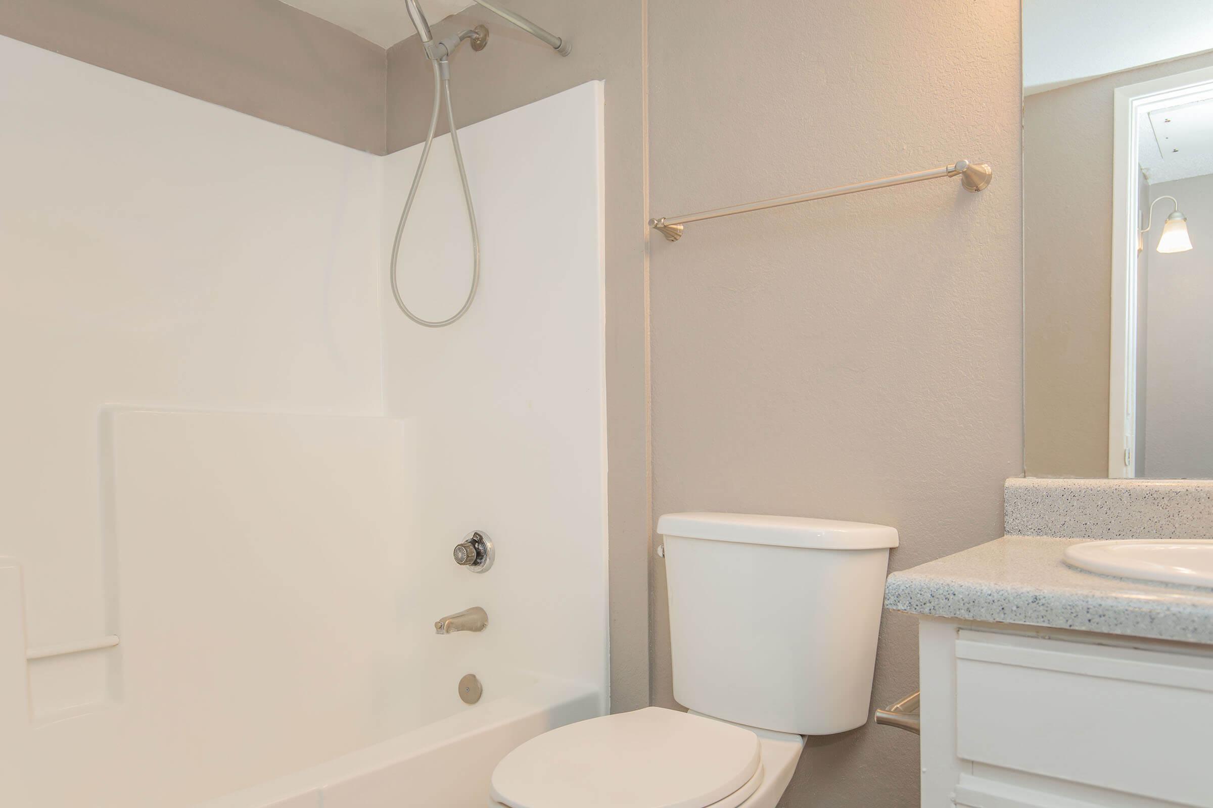 A clean bathroom featuring a white bathtub with a shower fixture, a white toilet, a countertop with a sink, and a large mirror. The walls are painted in light gray and beige tones, creating a modern and fresh appearance.