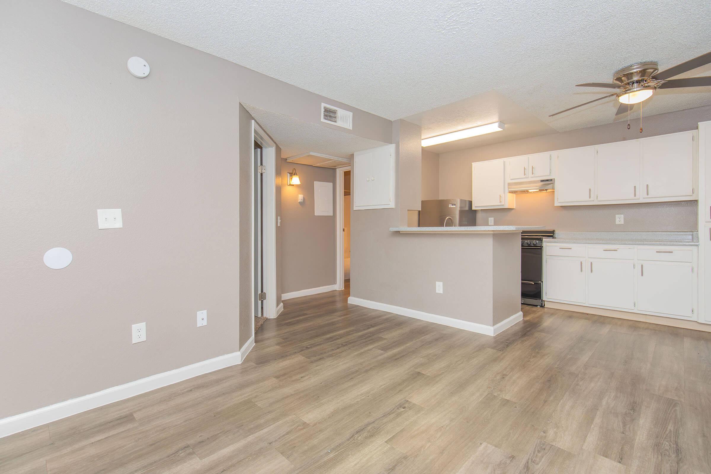 Interior view of a modern studio apartment featuring light gray walls, a wooden floor, and a ceiling fan. The kitchen includes white cabinets and appliances, while a small hallway leads to additional rooms. Natural light enters the space, creating a bright and inviting atmosphere.