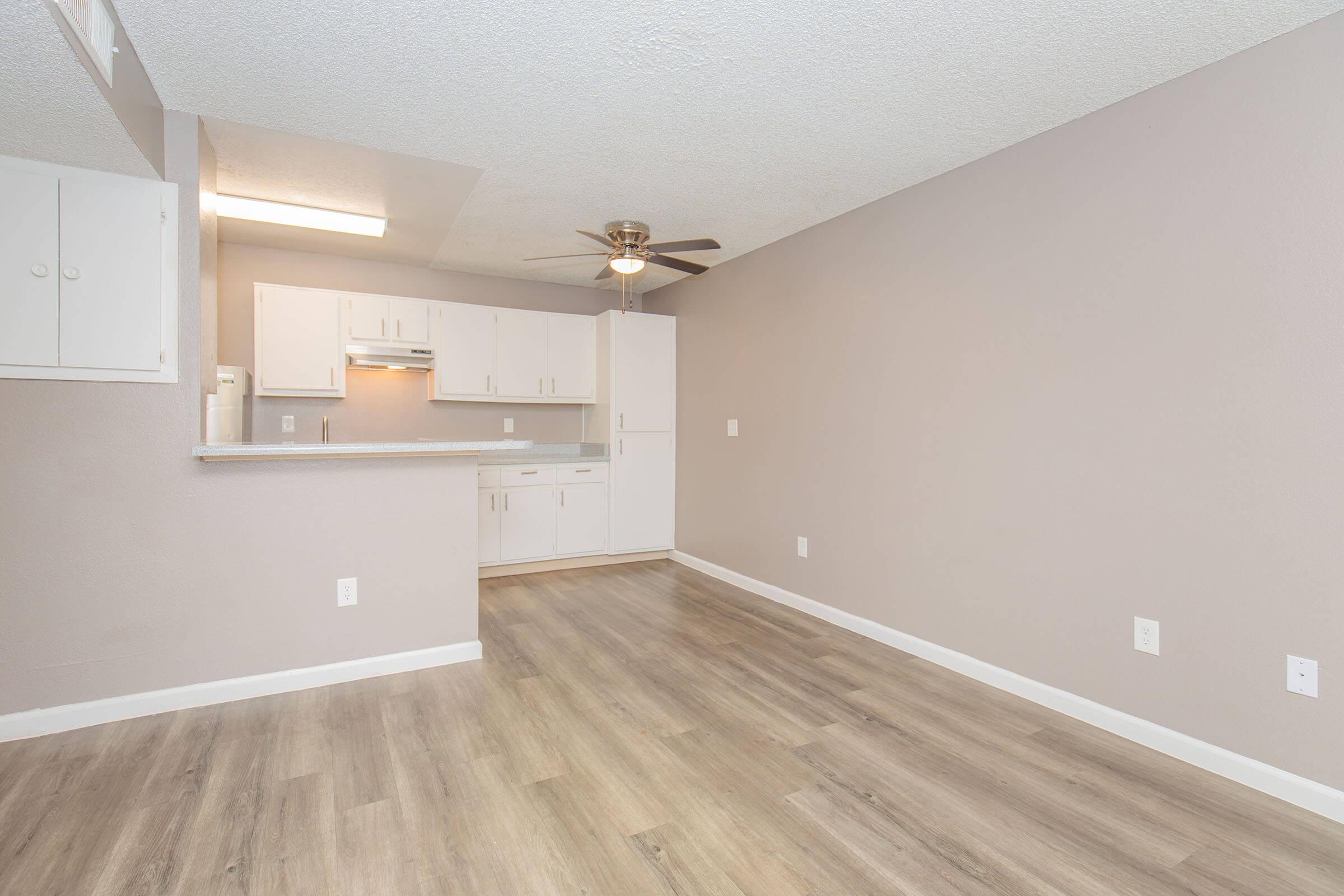 An interior view of a modern, open-concept living space featuring light-colored walls, a ceiling fan, and laminate flooring. The kitchen area is visible with white cabinetry and appliances, blending seamlessly into the living area, which is empty and well-lit.