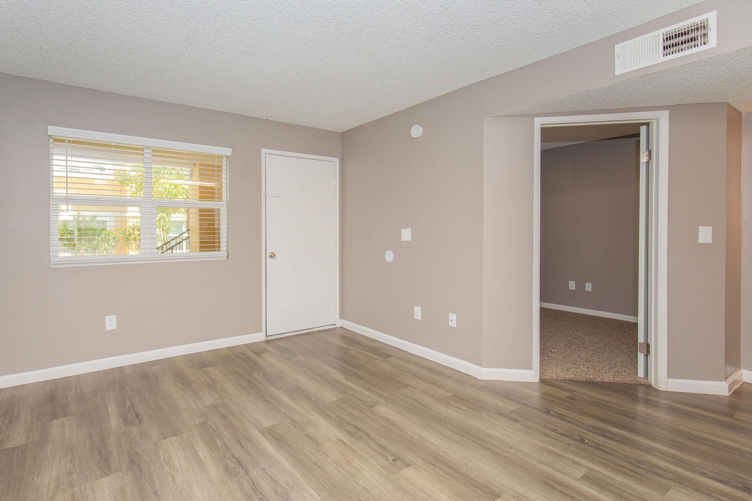 Empty living room space with light-colored walls and hardwood flooring. A window on the left allows natural light, and a white door leads outside. There's a doorway to another room on the right, and the overall atmosphere is bright and neutral.