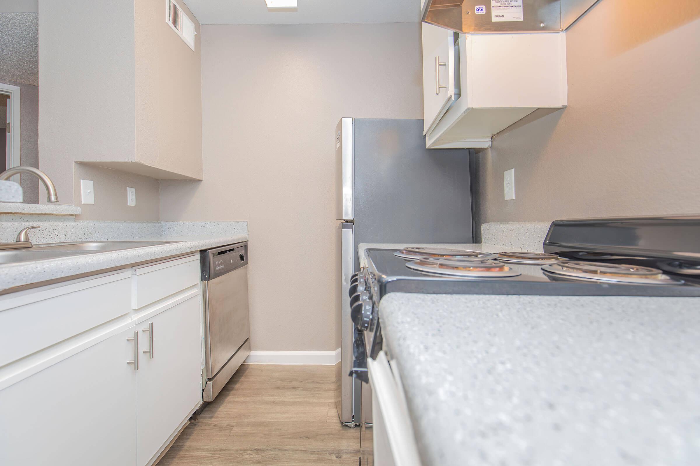 Kitchen interior featuring a countertop with a stove, a stainless steel dishwasher, and a refrigerator. The walls are painted in a light color, and there are cabinets above the countertop. The flooring is light brown, giving the space a modern look.