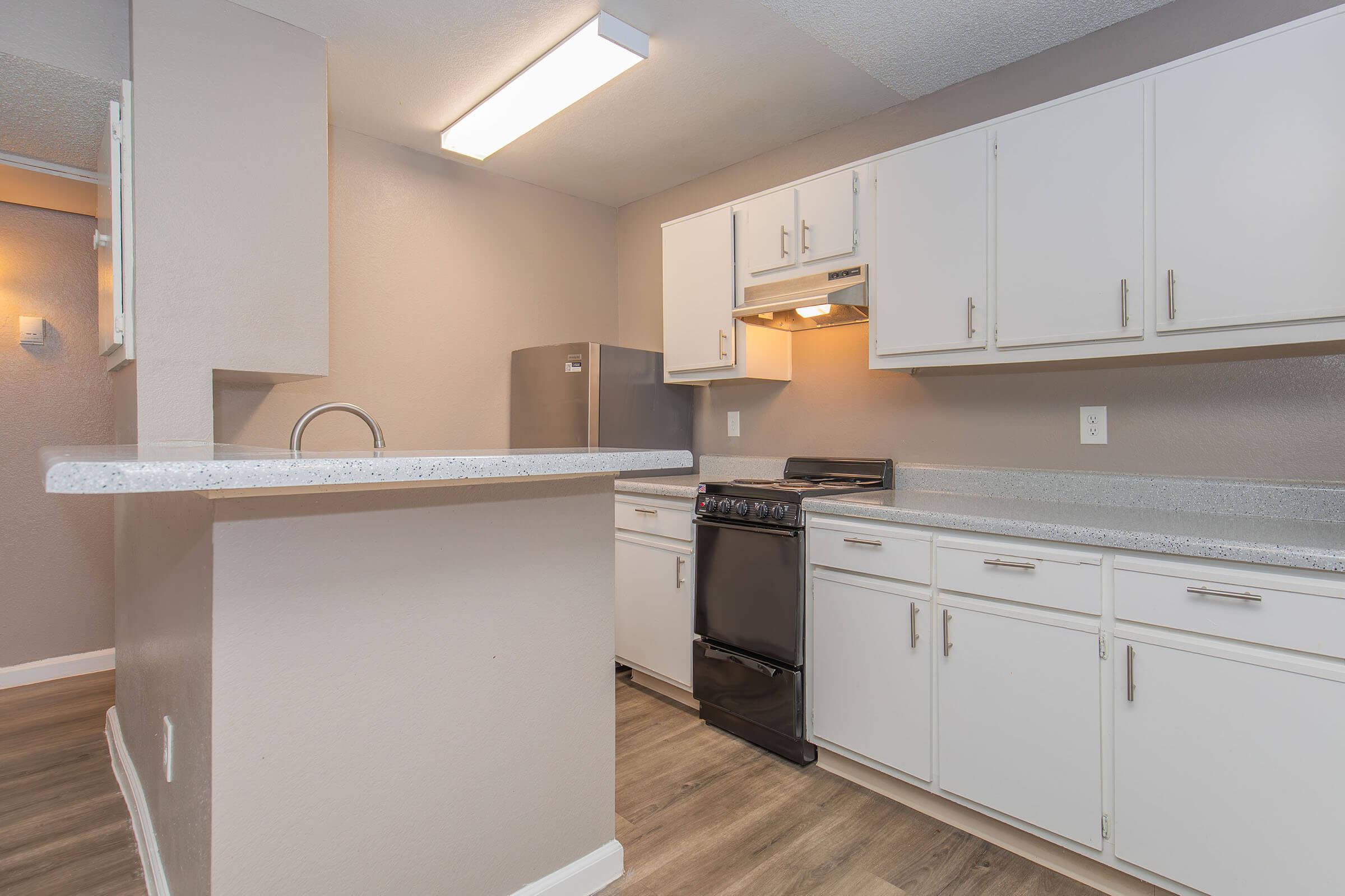 A modern kitchen featuring white cabinetry, stainless steel appliances, and a countertop with a built-in sink. The space includes a black stove and an open design that connects to a living area. Soft lighting enhances the neutral color scheme of the walls and flooring, creating a welcoming atmosphere.