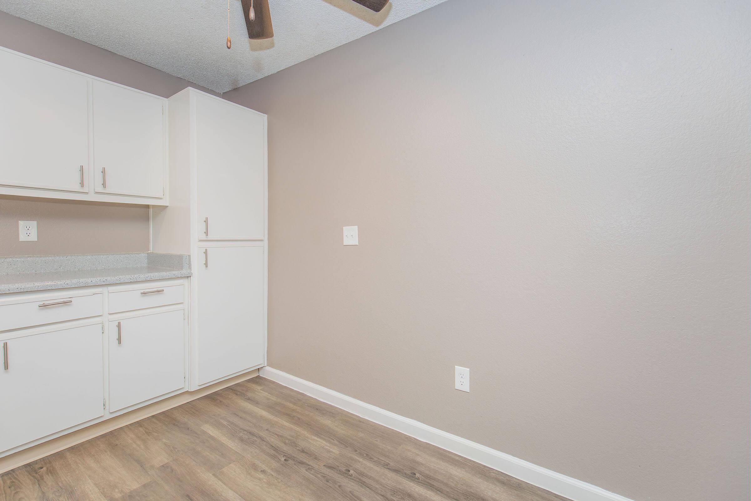 A clean, minimal kitchen space featuring white cabinetry, a light grey wall, and a wooden floor. There is a refrigerator and counter space, with a ceiling fan visible in the upper corner. The overall aesthetic is modern and uncluttered, inviting a sense of openness.